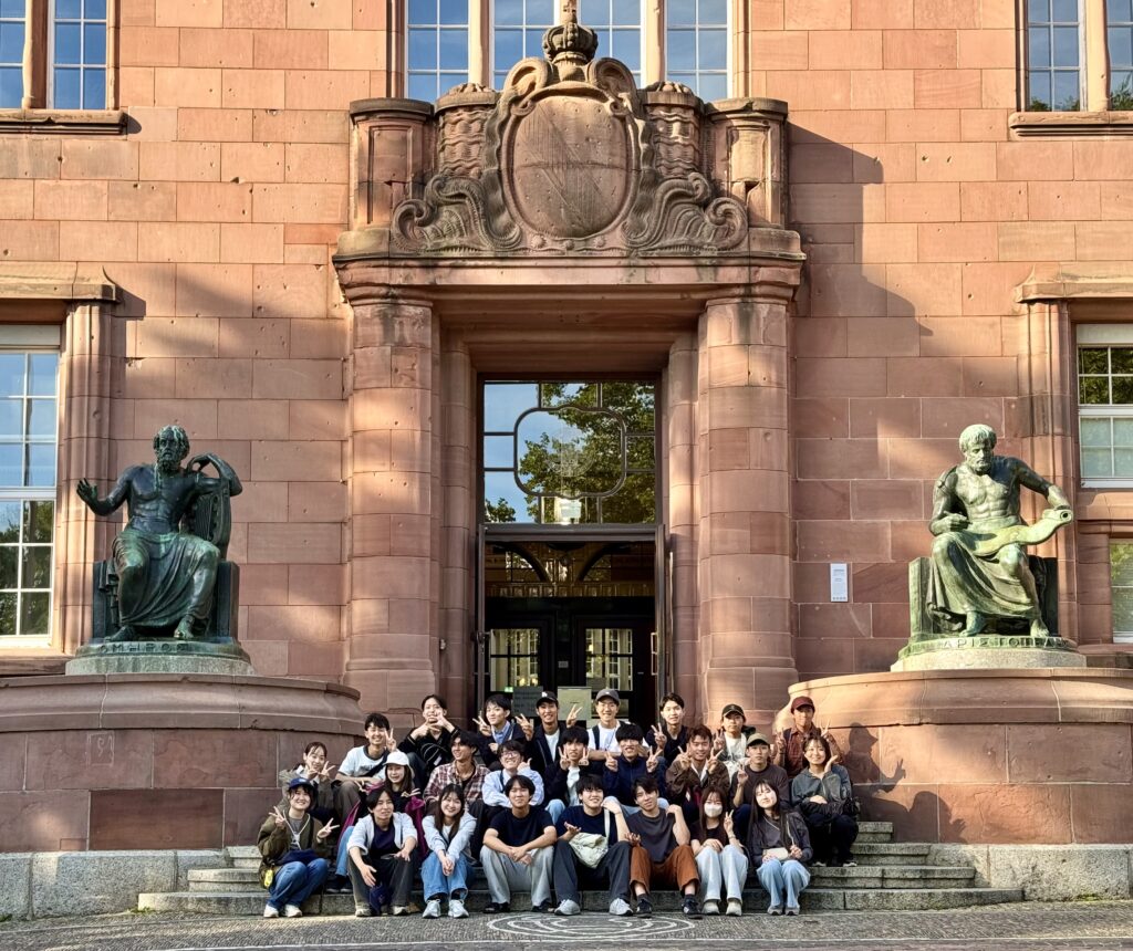 フライブルク大学の一号館を風景にした、ホメロス・アリストテレスの銅像の間に座っている2026年9月の研修生のグループ写真です。